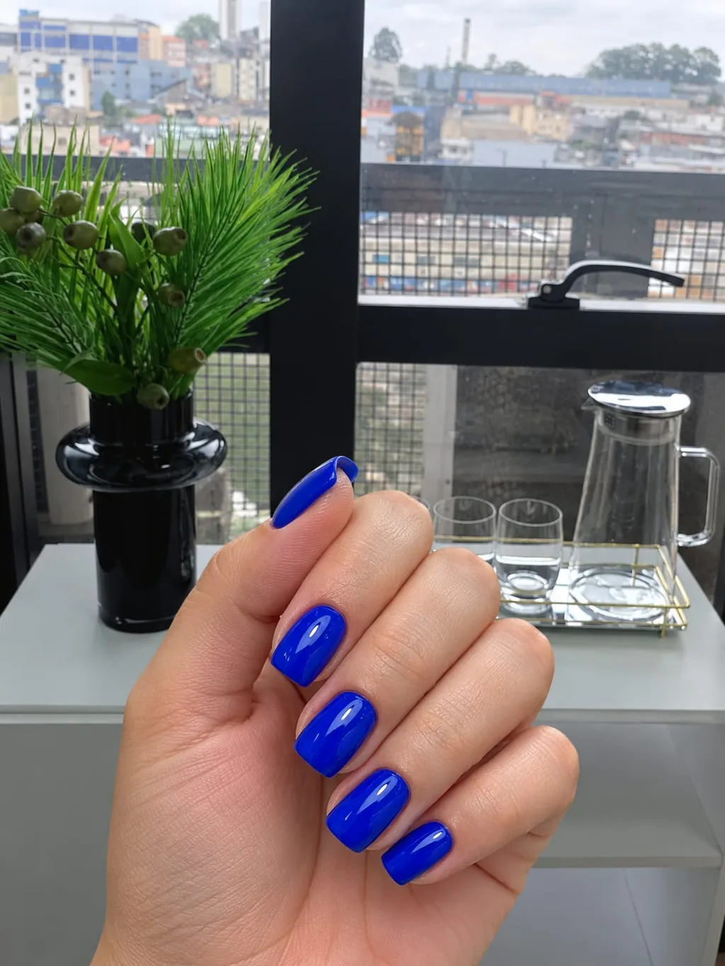 Unhas curtas quadradas com esmalte azul royal, mão em frente à janela