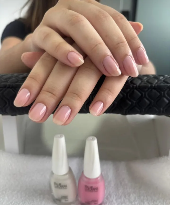 Unhas naturais com esmalte rosa nude, mãos sobrepostas e dois esmaltes embaixo