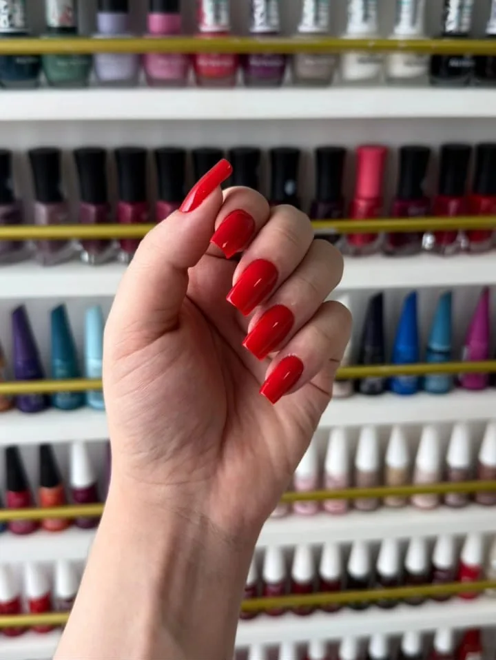 Mão com unhas vermelhas quadradas diante de uma prateleira de esmaltes